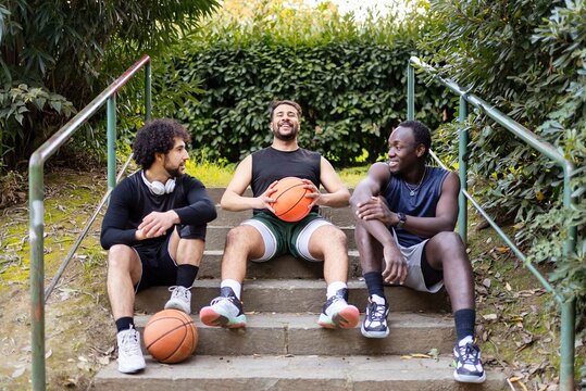 Three basketball players relax on a set of outdoor steps, laughing and chatting while holding basketballs, surrounded by greenery in a peaceful park setting