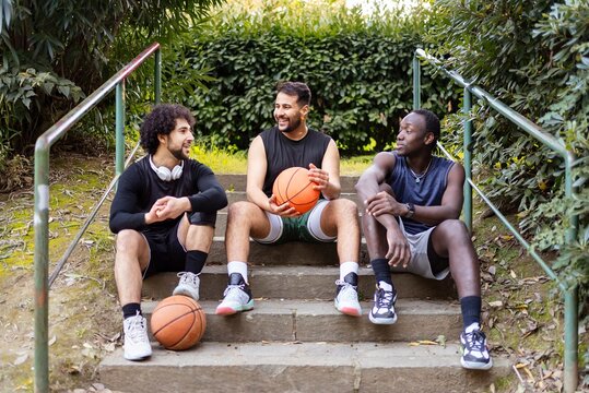Three basketball players relax on a set of outdoor steps, laughing and chatting while holding basketballs, surrounded by greenery in a peaceful park setting