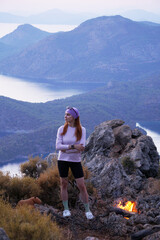 Naklejka premium A young girl stands by a campfire in the mountains with a beautiful sunset view