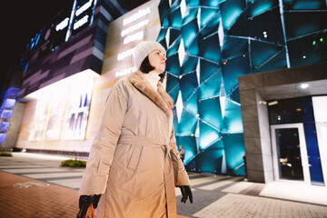 A woman in a light coat and a warm hat walks along a busy street in the evening city.