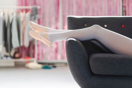 Legs of a ballerina wearing white tights and beige ballet shoes extended on a sleek gray sofa with a blurred background of clothing racks and pink decorations in a bright studio