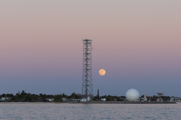A coastal scene at dusk with a full moon rising behind a tall metal tower and a white geodesic...