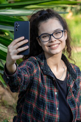 Young white girl in casual clothes takes a selfie with her mobile phone in a green park while smiling