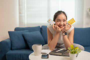 Young asian woman holding credit cards and smiling while sitting on a comfortable sofa in her...