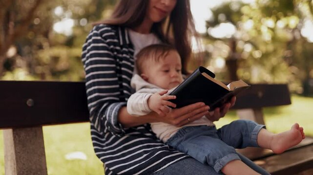 A young woman mother with a child in her arms read the bible together in the park outdoors. Mother teaches her son the word of God. Christian way of life, raising children, catechism