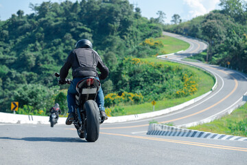 Obraz premium Motorcyclist rides along a scenic winding mountain road, surrounded by lush greenery and vibrant yellow flowers, under a clear blue sky.