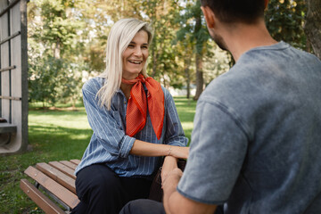 Fototapeta premium A couple sits on a park bench having a cheerful conversation on a sunny day. The woman smiles warmly, creating a relaxed and positive atmosphere in the lush green environment.