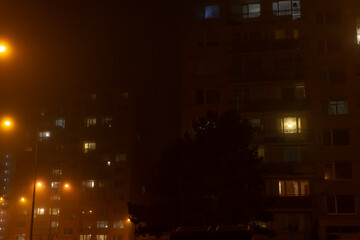 In a foggy urban setting at night, the scene is enriched by the soft glow of illuminated apartment windows and the warm light of streetlights, creating a mysterious yet soothing atmosphere