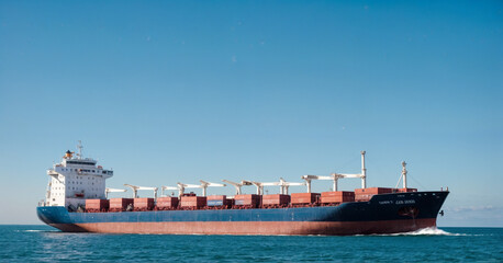 Fototapeta premium A large container ship navigating through the ocean under a clear blue sky, loaded with shipping containers, representing global trade, logistics, and maritime transportation.