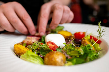 A close-up o hand garnishing a gourmet salad with fresh greens, vegetables, and a poached egg on a white plate. The vibrant colors and textures highlight the freshness and quality of the ingredients