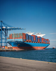 Fototapeta premium A large container ship navigating through the ocean under a clear blue sky, loaded with shipping containers, representing global trade, logistics, and maritime transportation.
