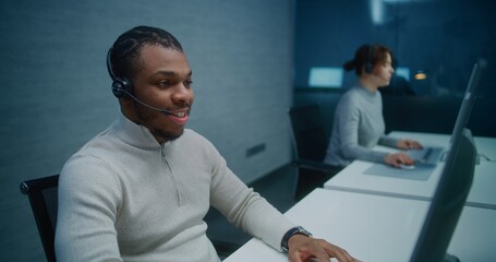 Call center: African American helpdesk specialist talks on headset with customer, works on...