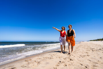 Two beautiful middle-aged women wearing one-piece bathing suits and sarongs walking and talking, one pointing something, on sandy beach in summertime during holidays. Front view.