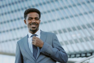 Fototapeta premium Portrait of confident businessman smiling while holding his tie in urban city setting showing modern architecture background