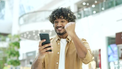 Happy young handsome man rejoices at the good news looking at the smartphone phone standing in shopping center mall store Joyful find sales discount online Surprised male celebrating victory indoor