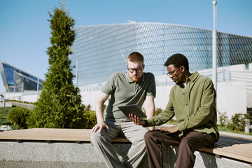 Two men discussing project on tablet while sitting on bench in modern urban area with glass architecture in background