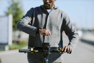 Person in casual attire checking smartwatch while standing on electric scooter, outdoors with blurred background suggesting movement and activity in urban area