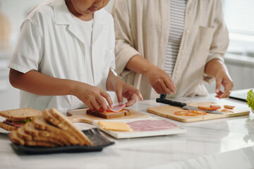 Two individuals are in kitchen, hands busy preparing ingredients for sandwiches. Fresh vegetables, cheese, and bread slices are laid out on countertop adding to culinary activity