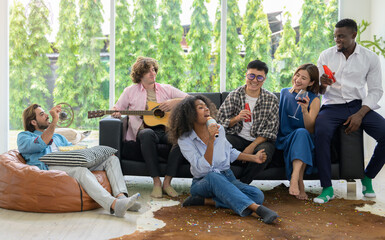 Group of diverse friends sitting on couch and enjoy drinking in living room while having party at home. Cheerful African woman singing a song while Caucasian man playing guitar at weekend..