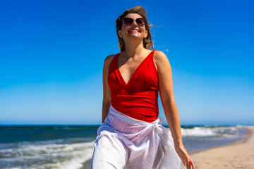Woman radiating happiness. Portrait of beautiful young girl in red swimsuit and white pareo strolling by sea on sandy beach on beautiful sunny day. Joy of life.