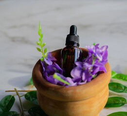 A mortar with purple flowers and an amber vial full of essential oil, on a marble surface. Close-up image