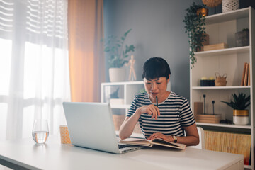 Asian woman working on laptop in home office with book and pen