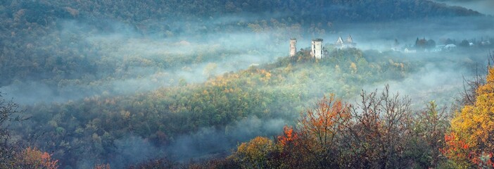 wide-angle panorama landscape with tales of morning fog between forests hills and ruins of medieval castle in autumn morning