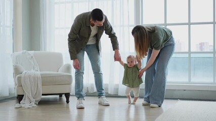 Baby development. Little infant girl making first steps at home. Happy mother father looking at toddler child walking in living room. Happy family at home. Mom dad supporting baby first steps