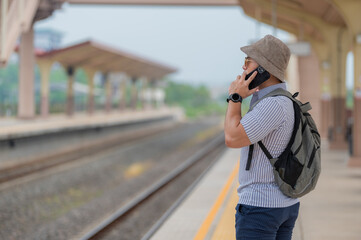 Fototapeta premium Handsome young man with a backpack uses the phone while standing near the railroad train on the platform,Tourism and travel in the summer. Vacations for the student.