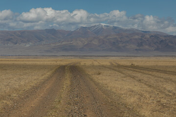 Naklejka premium Long curved country road running along the hilly steppe. Road lead to the mountains ridge in the distance. Golden autumn roadscape: endless expanses, emptiness, abandoned and deserted territory.