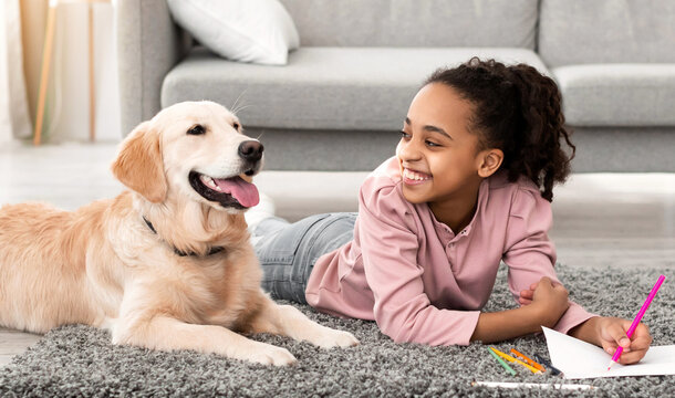 Children And Domestic Animal. Portrait of cheerful black girl lying on the floor carpet with her dog, drawing with colorful crayons, looking at golden retiever in living room, banner, panorama