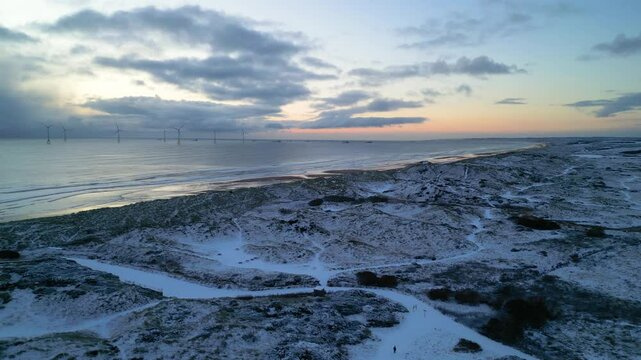 Person walks on fresh snow out to snow covered beach at sunrise. Drone strafes to reveal wind turbines and boats on horizon on clear morning. Aberdeen. 