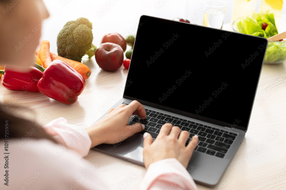 Aufkleber Over the shoulder view of young female using personal computer with black empty screen for mock up design, checking email or browsing internet sitting at dinner table in kitchen, searching recipe