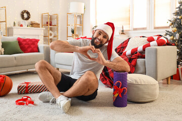 Obraz premium Young happy man in Santa hat with gift boxes and ball showing heart gesture at home decorated for Christmas