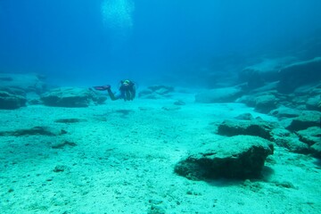 Fototapeta premium Scuba diver exploring underwater landscape with blue ocean. Deeper rocky seascape, clear sea and swimming underwater explorer. Travel picture, adventure scuba trip, being free in the sea. 