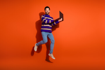 Full body photo of attractive young man jump hold netbook dressed stylish striped purple clothes isolated on orange color background