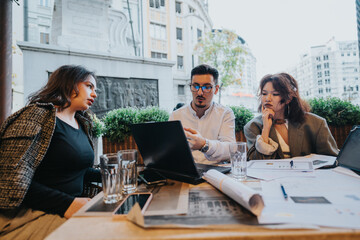 A group of diverse business people engaged in a meeting at a coffee bar, discussing and analyzing project ideas collaboratively. They are intensely focused on their laptops and documents.