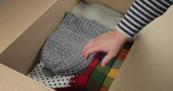 A woman puts warm sweaters, scarves, hats and long sleeves in a cardboard box. Content about storing, moving, sorting, mailing or donating clothes.