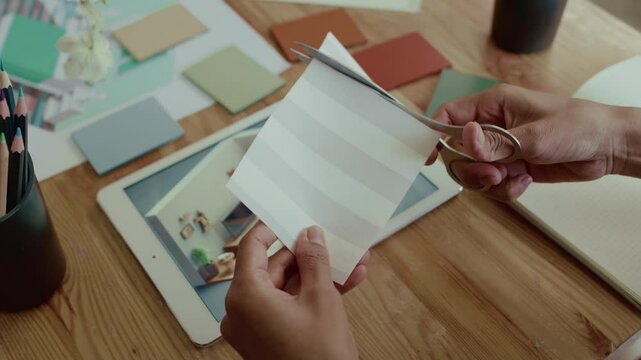 Close-up view of hands of unknown female designer cutting off part of decorative paper with scissors while planning interior for living room, digital tablet with 3D model on desk