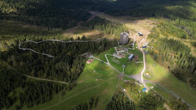 Aerial drone view of the Bachledka Treetop Walk and observation tower surrounded by dense mountain forest. Scenic tourist attraction with elevated wooden canopy walkway, ski lift infrastructure and pa