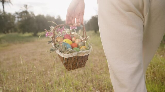 A Woman Walks Through a Field, Holding a Woven Basket Filled with Colorful Easter Eggs and Flowers. Slow motion.