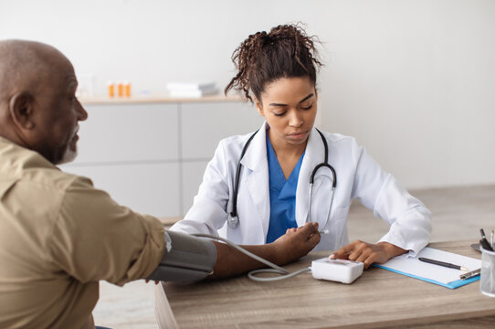 Young Focused African American Woman Examining Older Patient Measuring Arterial Blood Pressure With Modern Tonometer Using Medical Device Sitting At Table. Hypertension Health Problem Concept