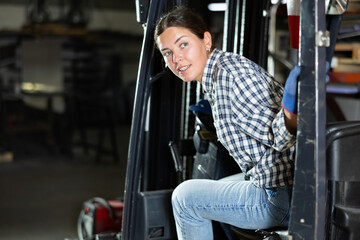 Young female worker in uniform posing while sitting in forklift in metallurgical workshop