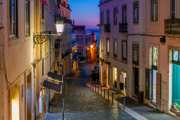 View of the Tagus River and colorful twilight blue hour sky at evening sunset from a narrow hillside cobblestone street in the medieval Alfama district of Lisbon, Portugal.