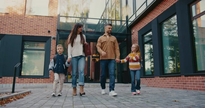 Front camera view of parents taking their kids from school. Caucasian man talking with woman while walking together in same direction. Children happy finishing difficult study day.