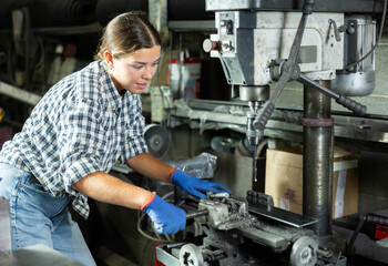 Skilled young female factory worker, dressed in plaid shirt and protective gloves, working on drilling machine in metalworking workshop, performing holes in flat steel part..