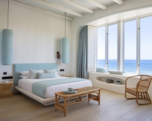 Fototapeta premium Modern coastal bedroom with whitewashed walls, light blue accents, rattan furniture, and large windows offering ocean views, creating a serene and beachy vibe.