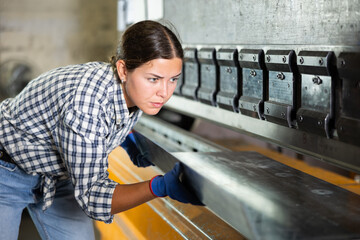 Young female worker in plaid shirt and gloves operating press brake in industrial workshop,...