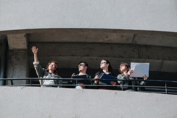 A diverse group of business people stands on a building balcony, holding documents and discussing...