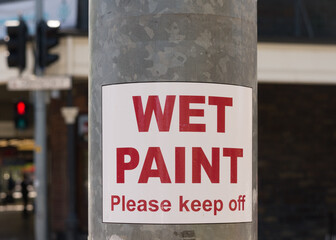 Metal post with signage indicating wet paint with out of focus urban background
